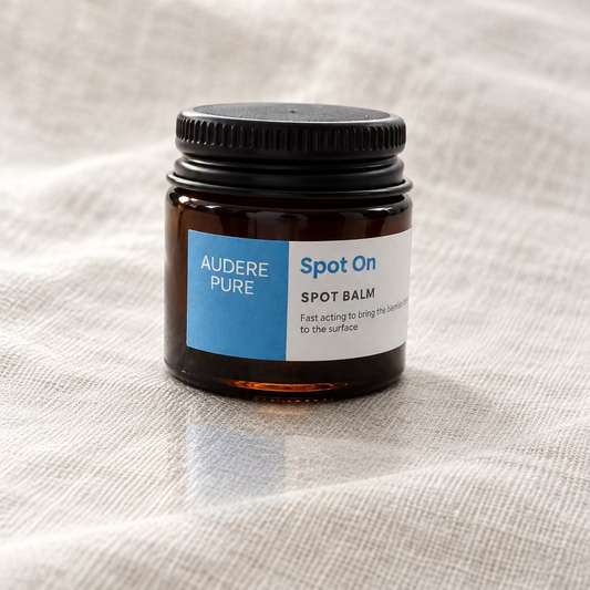 A small amber glass jar of Audere Pure Spot On Spot Balm with a black lid and a blue and white label, sitting on a white surface.